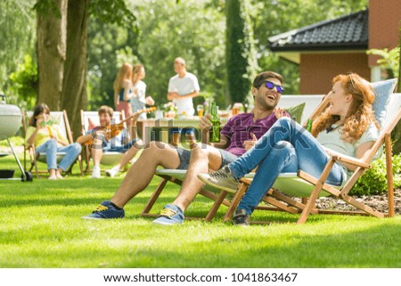 Young friends having fun at backyard barbecue party, drinking beer, talking and playing guitar on hot summer day, focus on the couple laughing sitting on deck chairs Royalty-Free Stock Photo #1041863467