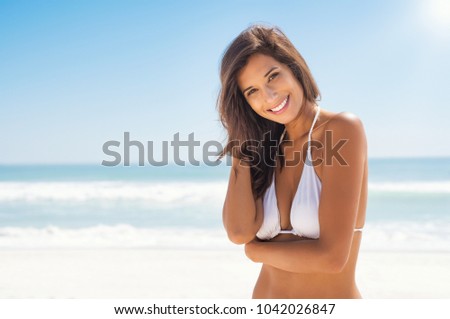 Beautiful young woman at beach looking at camera. Happy latin girl in white bikini smiling. Portrait of young tanned woman relaxing on beach with copy space and sea in background.