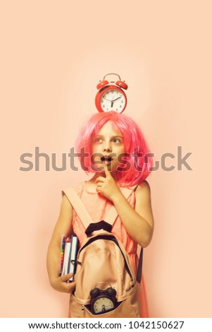 Girl with wig in pink dress with backpack. Pupil with thoughtful face expression, isolated on light pink background. Back to school and learning concept. Kid with alarm clock on head and books in hand