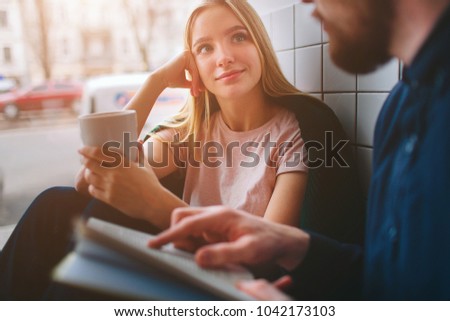 A girl sitting in cafe near big window and looking to her boyfriend. Se is listening to him very careful while he is reading a book for her. Close up. Cut view.