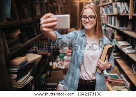 A cute picture of a blonde girl taking selfie. She is looking to the phone and smiling. This girls is in a big public library.