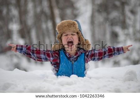 A little boy is standing in a deep snow bank after a snow storm. He has a furry winter hat with ear flaps and a plaid shirt with snow pants. The child thinks the deep snow is fun and is laughing. 