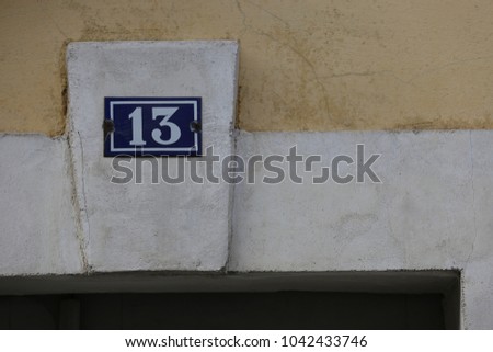 Close up outdoor view of the number 13 written in white on a blue metallic rectangular plate. Symbol fixed above a door to indicate the address. Wall made of stones with white decorative elements. 