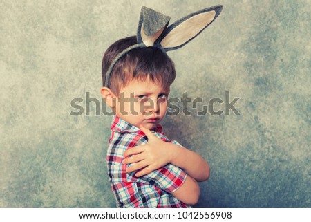 A funny, grumpy little boy dressed in a red checkered shirt and an old-fashioned hat, has a rabbit's ears on his head. Copy space.