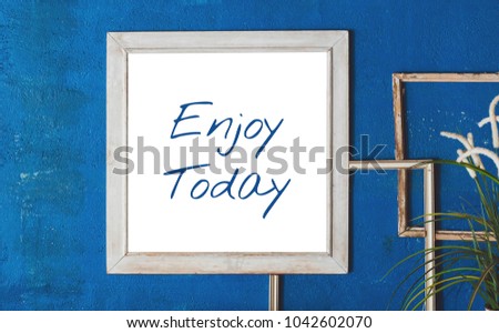 White wooden frame with quote "Enjoy Today" on blue wall background.