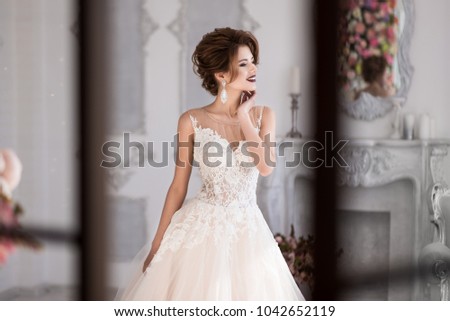 Morning of the bride. A beautiful bride is standing in a lace wedding dress. Wedding hairstyle and makeup. Hand at the face. Photo through the window.