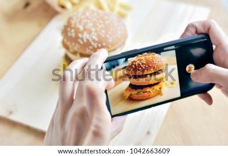 Treandy woman using her smartphone taking the fresh tasty big burger lunch photograph for sharing on her social community.Daily routine before eating every meal.Burgers are an American icon.