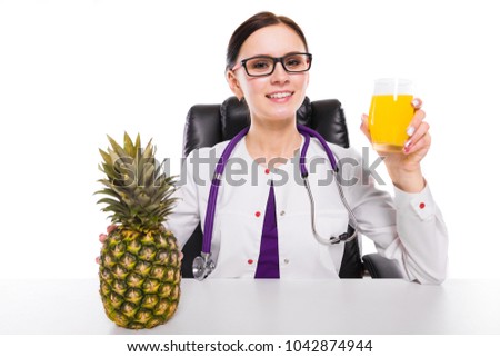 Female nutritionist sitting in her working place showing and offering glass of pineapple fresh juice holding pineapple in her hand on white background