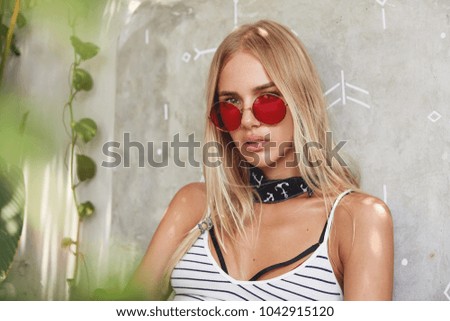 People, beauty and style concept. Beautiful young blonde woman wears red trendy shades, striped t shirt, has serious expression, enjoys calm atmosphere at home. Relaxed hipster girl models indoor