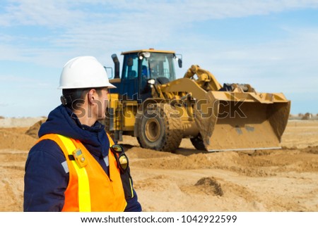 During leveling construction site young signalman, spotter gives visible signals with radio to heavy equipment, forklift and grader. Perfect background of construction operations.