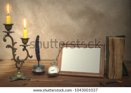Empty photo frame with copy space, book and burning candle in vintage candlestick and feather pen with inkpot and pocket watch on retro wooden table background. Education. Back to school. Memoirs.