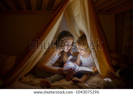 Two girls read fairy tales at dark night under a blanket by the light of a small lamp Royalty-Free Stock Photo #1043075296