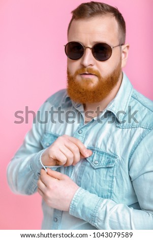 smiling red-haired beard man in sunglasses and denim shirt on pink background.