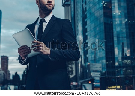 This is my city. Night time image of confident young man in full suit holding digital tablet and looking away while standing outdoors with cityscape in the background
