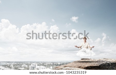 Young man in white clothing keeping eyes closed and looking concentrated while meditating on cloud in the air with city view on background.