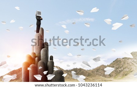 Businessman in suit with monitor instead of head keeping arms crossed while standing on the top of stone columns among flying paper planes with beautiful landscape on background. 3D rendering.
