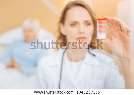 New pills. Smart experienced professional doctor looking at the box with new pills while her ill tired patient staying in bed behind her back