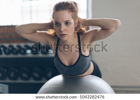Fit young woman doing pilates exercises in a gym balancing over a gym ball with her hands behind her head toning and strengthening her muscles