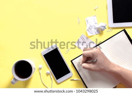 Man is going to write something on his notebook. Flat lay home office workspace with tablet, diary, mobile and white accessories in vintage pastel colored tone on yellow background with copy space.