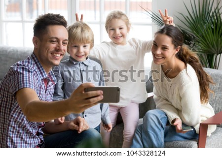 Smiling girl making mother and brother bunny ears while father taking happy family selfie, playful adopted children having fun posing for funny photo on smartphone with loving mom and dad together