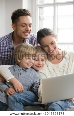 Happy family with adopted kids having fun using laptop together sitting on sofa, parents and son daughter relaxing at home with computer, smiling couple with children watching video online, vertical