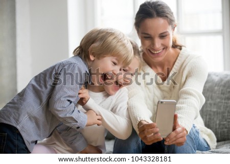 Smiling mother taking selfie with cute kids on smartphone, happy young mom laughing making photo with little son and daughter at home, single mommy and adopted children playing having fun with phone