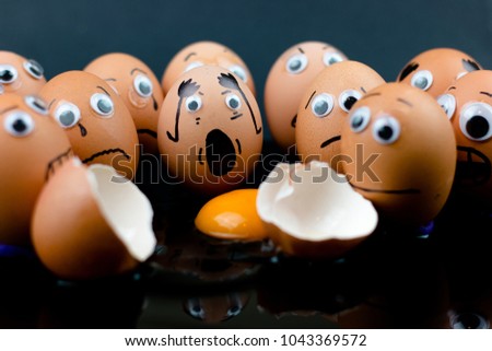 Fun concept: raw eggs with googly eyes and drawn features are in shock and sad as they witness another egg broken in two with the white and yolk in front of them; dark background.