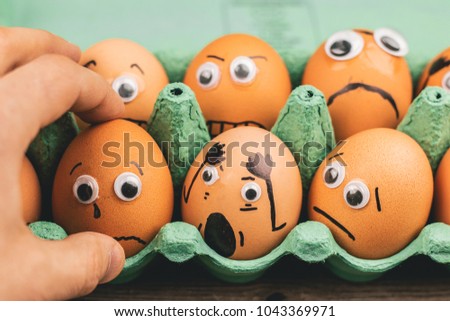 Fun concept: raw eggs with googly eyes and drawn features are in shock while sit in a green carton box and are being picked up by a hand