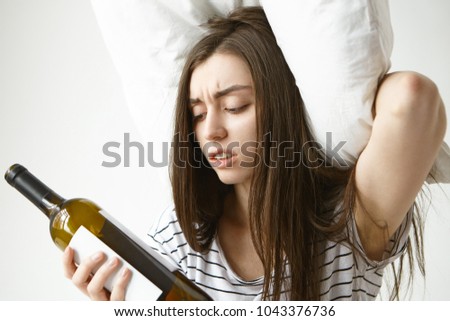 Picture of stressed frowning young Caucasian woman in striped pajamas holding pillow on her head and looking at empty bottle, feeling desperate as wine is over, suffering from hangover after party