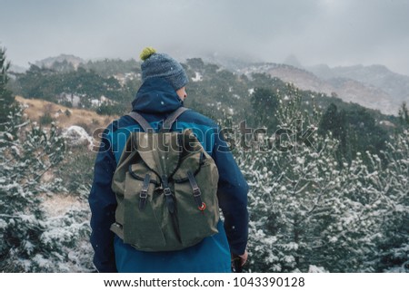 Man with backpack hiking in mountains. Backpacker looking into the distance. Cold weather, snow on hills and trees. Winter trekking. Back view.
