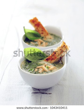 Smooth green broccoli soup in two white bowls standing on white table. Garnished with some pine nuts and basil leaves. Served with cheese croutons. 