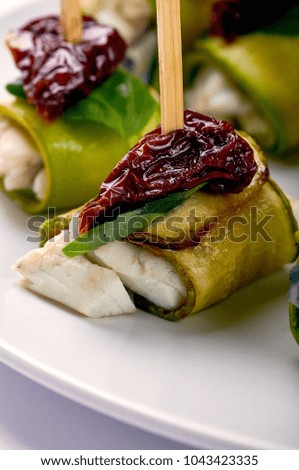 An unusual delicious appetizer from the chef. Roll with white fish, zucchini, dried tomato, prune on skewers. The food is decorated with a leaf of greens, basil, fresh spinach.