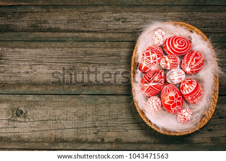 Red easter eggs with folk Ukrainian pattern lay on white feather in the basket which stand on vintage wooden board. Top view. Ukrainian traditional eggs pisanka and krashanka