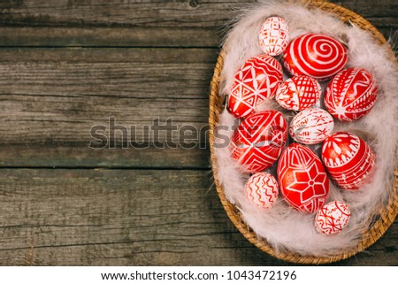 Red easter eggs with folk white pattern lay on white feather in the basket which stand on vintage wooden board. Top view. Ukrainian traditional eggs pisanka and krashanka