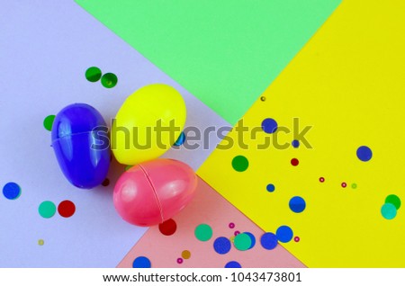 Three plastic easter eggs in blue, green, yellow and pink, with confetti on a geometric mutli-colored background made of rough textured paper. Studio lighting is reflected in eggs.