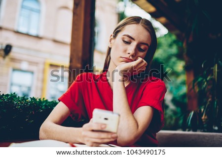 Sad hipster girl browsing boring websites on smartphone during leisure time in coffee shop outdoors.Dispirited female blogger reading repetitious news spending recreation time on social networks