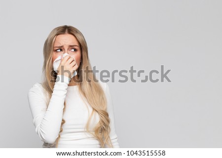 Studio portrait of unhealthy cute blonde female in white top with napkin blowing nose, looks to the source of the allergy, place for advertising/Rhinitis, cold, allergy concept/ Royalty-Free Stock Photo #1043515558