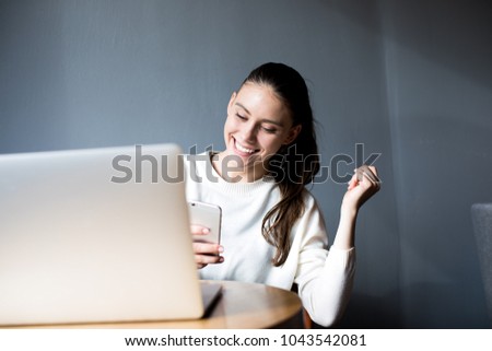 Happy woman writer received good news in text message on mobile phone during work on laptop computer. Smiling female student reading positive sms on her cell telephone, sitting with net-book in cafe Royalty-Free Stock Photo #1043542081
