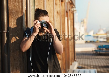 Capturing every moment of life. Outdoor shot of young stylish photographer looking through vintage camera, taking shots of harbour, yachts and seashore, working as freelance cameraman