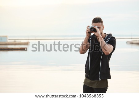 Stay where you are, this photo is amazing. Portrait of creative good-looking freelance photographer looking through camera while taking shots of nature and people, standing near seashore