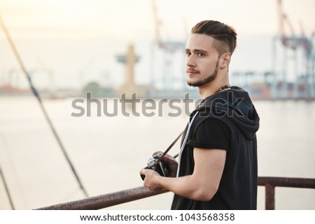 Sea attracts this photographer. Outdoor portrait of attractive young guy standing in harbour, enjoying looking at sea while holding camera, searching for good location to take photo, looking aside