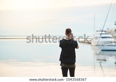 Sea takes your breath awat. Back view shot of stylish young european guy in trendy clothes standing on seashore taking photo of sea and beautiful yacht on smartphone, being photojournalist or amateur