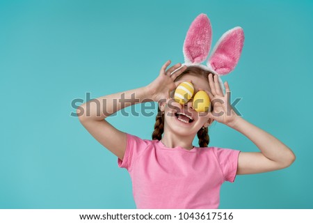 Cute little child wearing bunny ears on Easter day. Girl holding basket with painted eggs on light blue background. Royalty-Free Stock Photo #1043617516