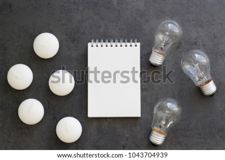 Notebook with candles and three lamp bulbs on the grey concrete background. Invention of electricity, light consumption concept. Top view, copy space, flat lay, mock up