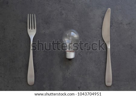 Light bulb, fork and knife on the grey concrete background. Electricity consumption, intellectual property concept. Top view, flat lay, copy space