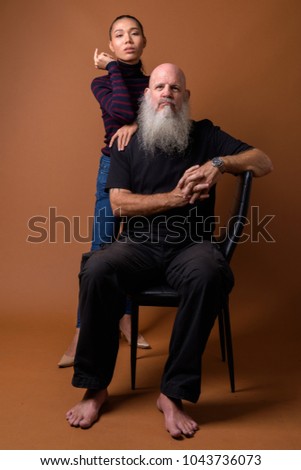 Studio shot of mature bearded bald man and young beautiful Asian transgender woman against brown background