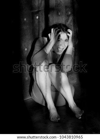 Black and white portrait of an emotional and screaming  woman in a dress, looking frightened at the light grasping her hair with her hands sitting on a stone floor against a rusty wall. Fear and Panic