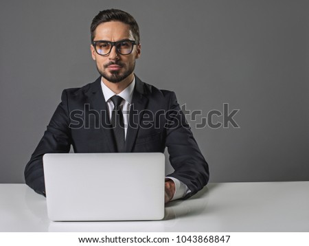 Waist up portrait of concentrated young man sitting at the white table with computer. He is looking at camera with confident look. Copy space in right side