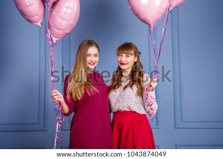 two young beautiful girls in red dresses stand on a blue background and hold in their hands the balls of the heart