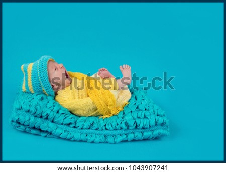 Baby boy is having his first photo shoot and he is yawning. He is dressed in yellow and blue clothes and is wearing a hat in the same colours.